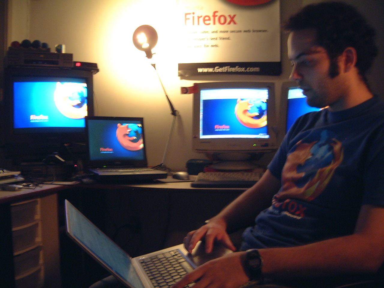 A person uses a laptop beside several older computers displaying Firefox branding, with a Firefox poster on the wall.