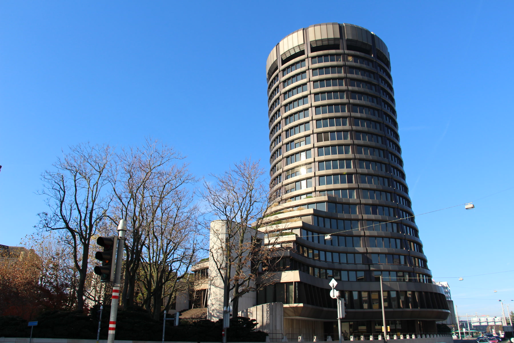 The round concrete tower of the Bank for International Settlements on a Basel street, with the Rhine and neighboring buildings beside it.