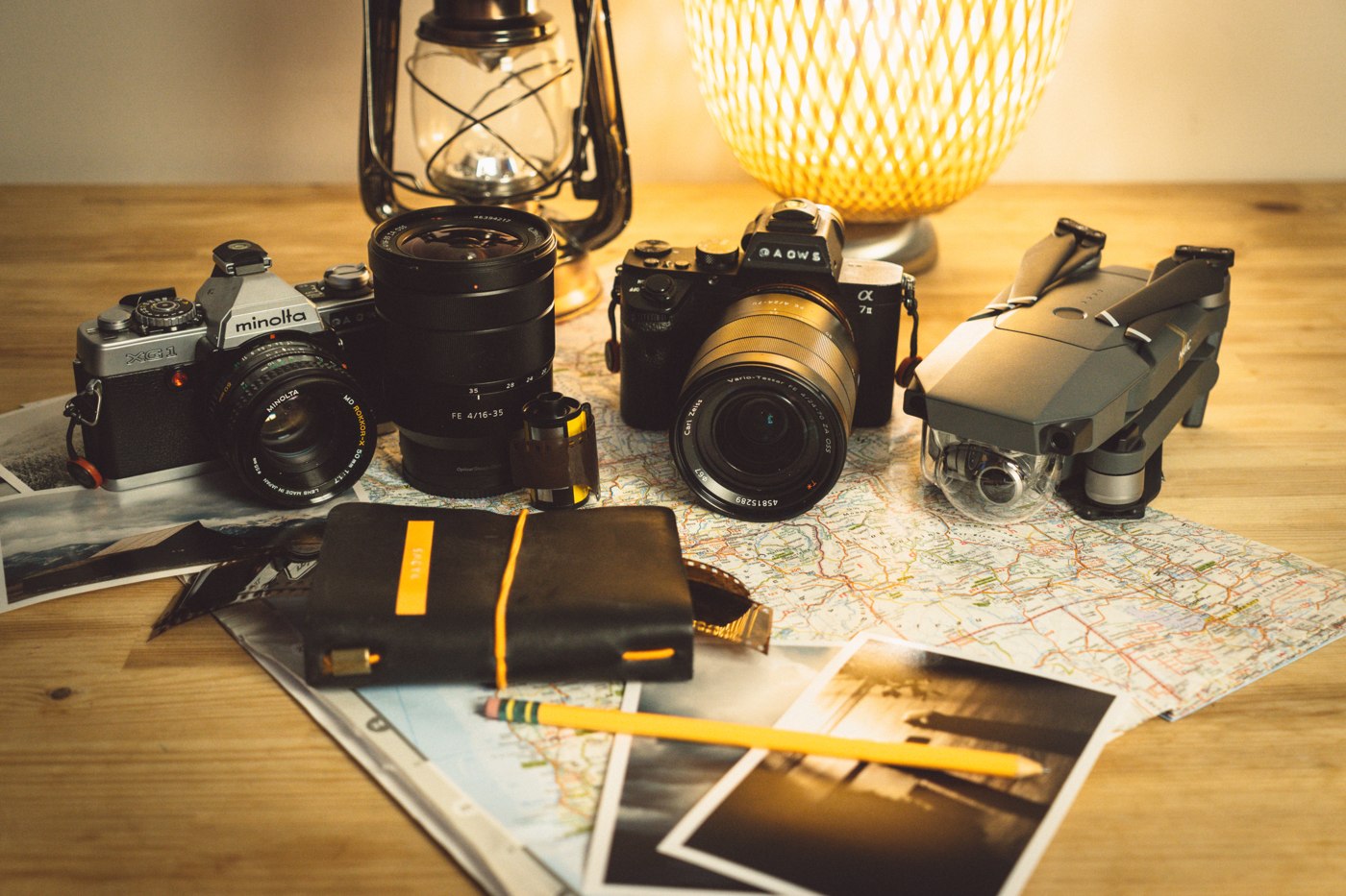 Two cameras, a folded drone, a lens, printed photographs and a notebook arranged on a map on a wooden table.