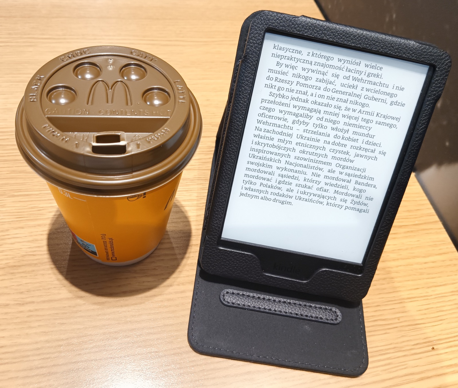 A Kindle in a black folding case displays a page of Polish text beside a takeaway coffee cup on a wooden table.
