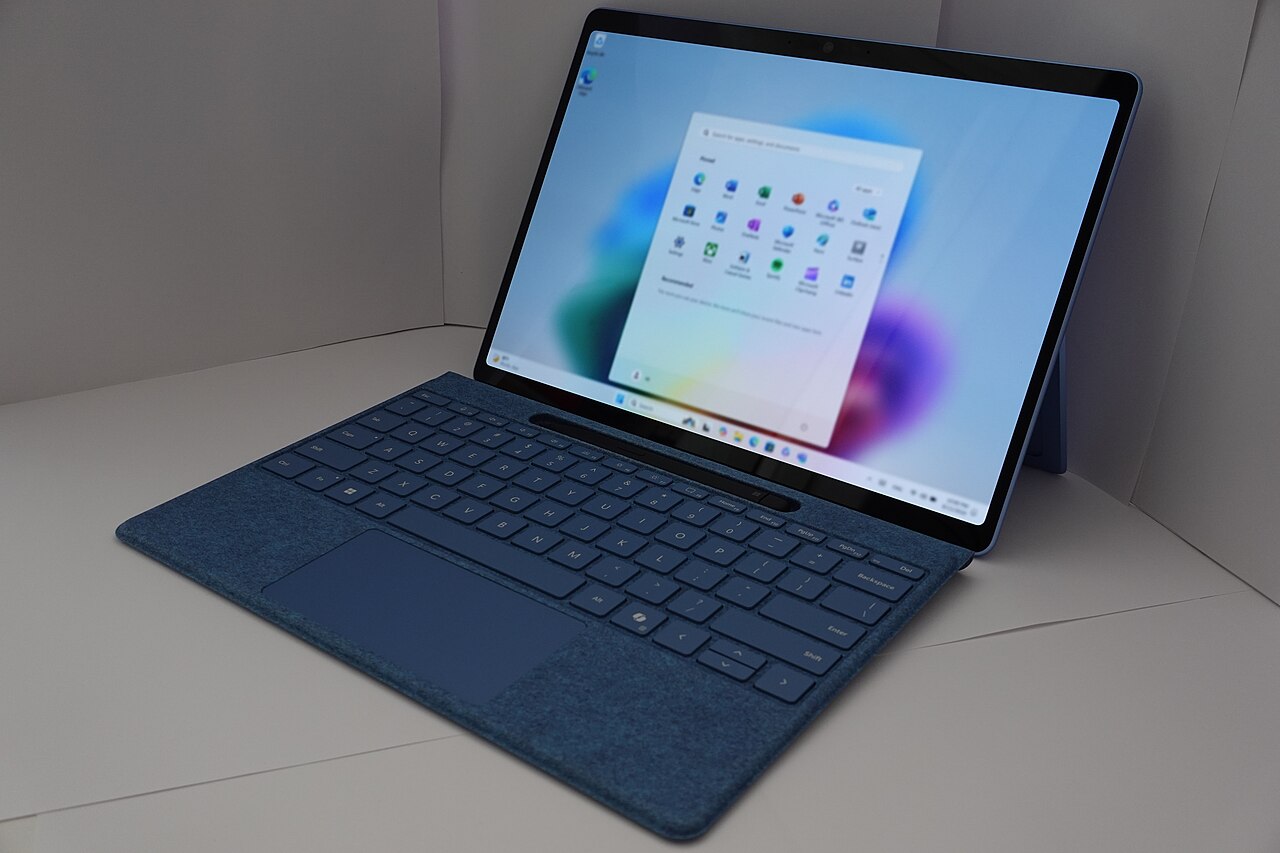 A Microsoft Surface computer with a blue keyboard cover and the Windows 11 Start menu on its screen.